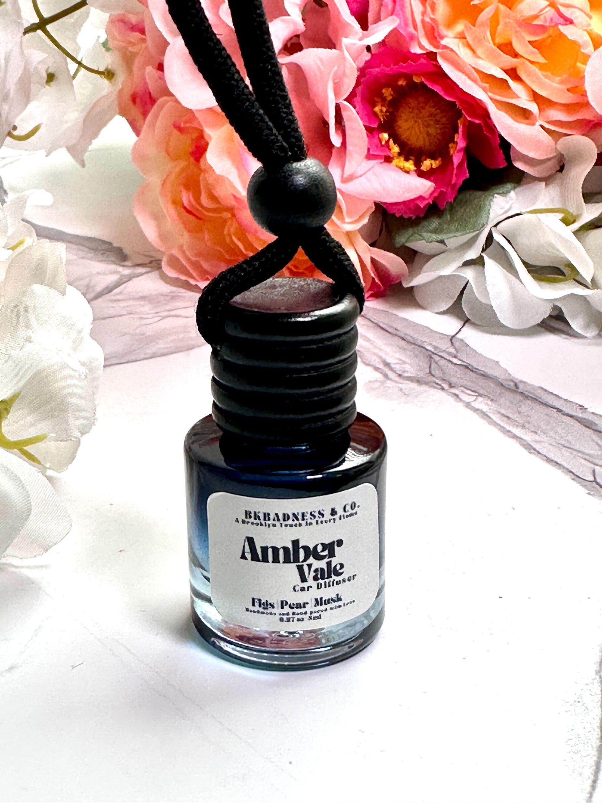 Amber Vale car air freshener bottle with black cap on a white surface with flowers in the background
